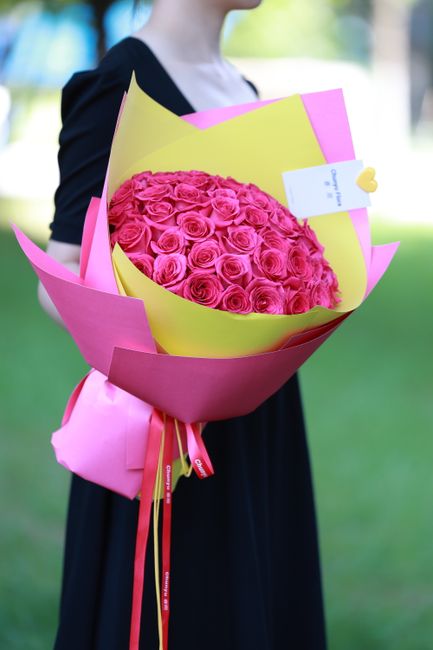 A person dressed in a dark dress is holding a large bouquet of fresh pink roses arranged in a tight, rounded cluster. The bouquet is wrapped in vibrant yellow and pink paper, with the pink wrapping fe