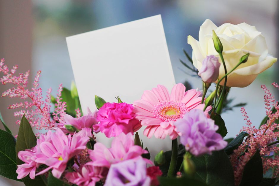 A close-up image of a floral arrangement featuring a variety of fresh flowers including white roses, pink gerbera daisies, purple lisianthus, and pink astilbe, all with vibrant, healthy petals and gre