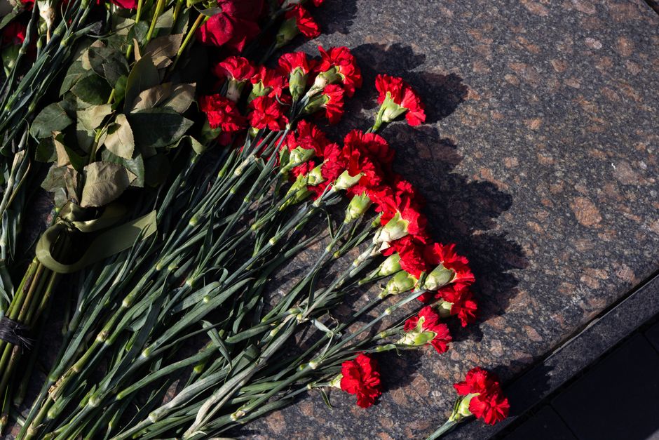 A close-up view of a bouquet of fresh red carnations with ruffled petals and long green stems arranged on a dark granite surface. Part of the bouquet is wrapped with dark green paper and tied with a m