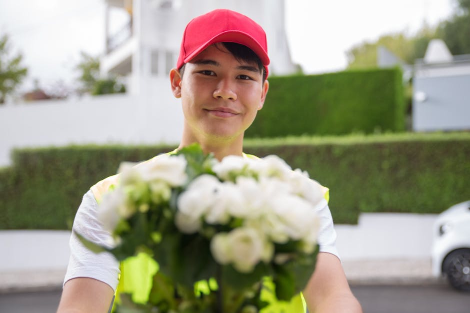 A delivery person wearing a pink cap and a light-colored polo shirt is holding a bouquet of fresh white flowers with green foliage, offering it towards the camera. The bouquet is wrapped in transparen