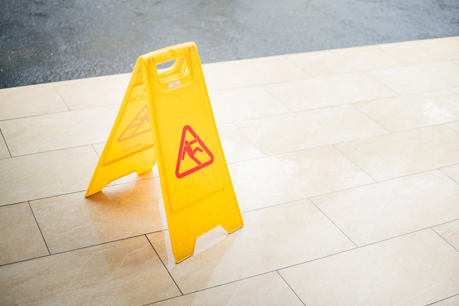A bright yellow, freestanding warning sign with red hazard symbols positioned on a beige tiled floor, indicating a wet or slippery surface. The sign is unfolded and upright, with faint shadows cast by
