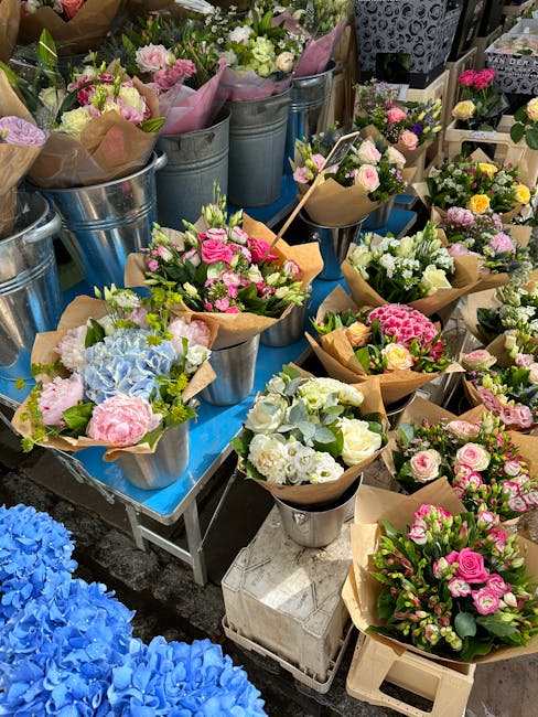 A variety of floral arrangements and bouquets displayed on a blue table outside a flower shop, featuring fresh flowers such as pink roses, white roses, light blue hydrangeas, and other colorful blooms