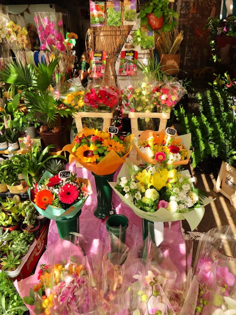 A vibrant display of floral arrangements in a flower shop, featuring bouquets of fresh flowers wrapped in colorful paper and leaves. Prominent flowers include bright pink gerbera daisies, yellow sunfl